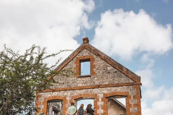Three people looking into the roofless brick and coral-stone shell of an old plantation house, against a bright sky