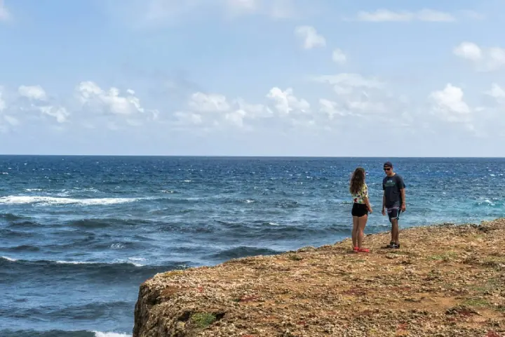 Two people standing at the edge of a low limestone cliff looking out at open ocean and breaking surf