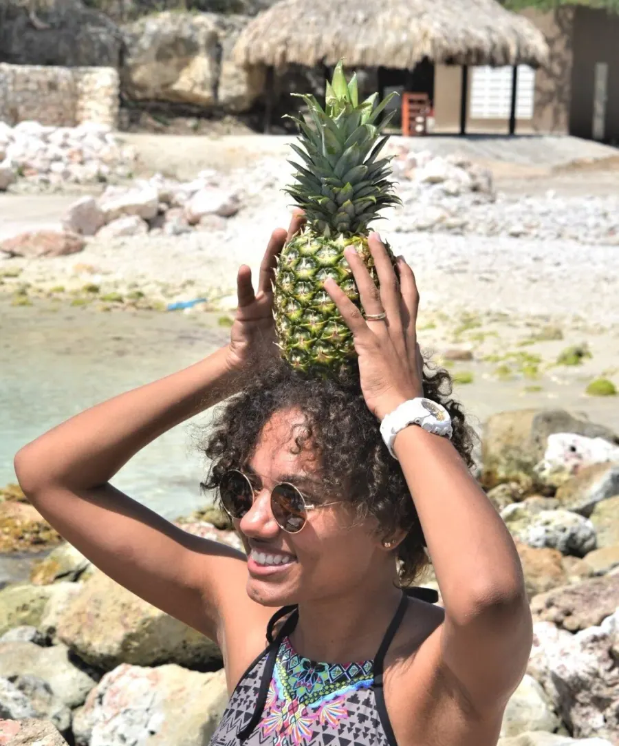 A woman in sunglasses laughing on a rocky beach, balancing a pineapple on her head