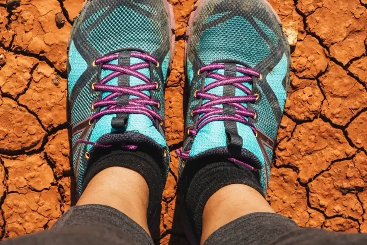 Looking down at a pair of worn turquoise hiking shoes standing on dry, cracked red earth